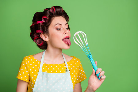 One small bite. Photo of charming pretty young lady roller hairdo stick out tongue lick dough baking cake hold plastic whisk wear dotted apron shirt isolated green color backgroundの写真素材