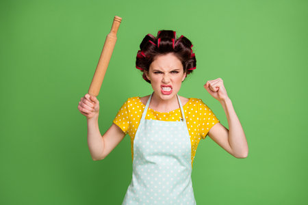 Photo of crazy evil furious lady curlers hairdo housewife hold rolling pin angry yell raise fist hate people step her kitchen wear pinup dotted dress apron isolated green color backgroundの写真素材