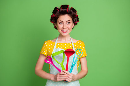 Photo of positive cheerful girl hold spatula spoon fork potato masher whisk enjoy prepare recipe wear good look clothes isolated over green color backgroundの写真素材