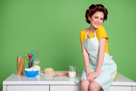 Photo of pretty attractive young lady sitting table improvisation kitchen curlers on hair charming housewife wear pinup clothes dotted dress apron isolated green color backgroundの写真素材