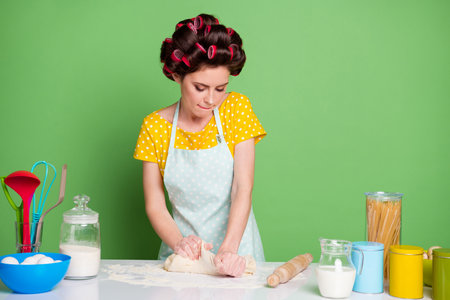 Photo of focused girl knead fresh pie cake dough table prepare home made dish wear yellow dotted t-shirt hair rollers isolated over green color backgroundの写真素材