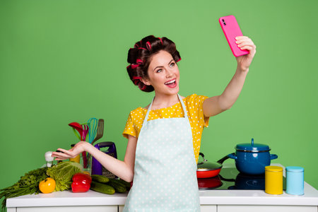 Photo chef girl influencer use cellphone make selfie hold hand demonstrate table fresh organic pepper ingredients wear yellow dotted t-shirt hair rollers isolated green color backgroundの写真素材