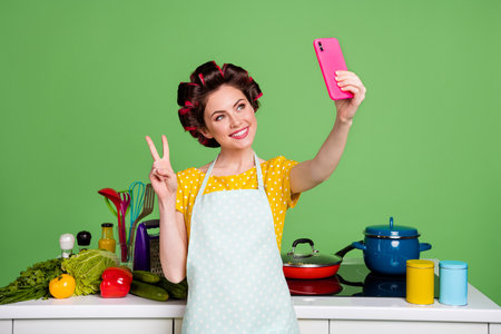 Photo positive girl prepare fresh eco supper use smartphone make selfie v-sign blogging ecological dinner wear yellow dotted t-shirt hair rollers isolated green color backgroundの写真素材