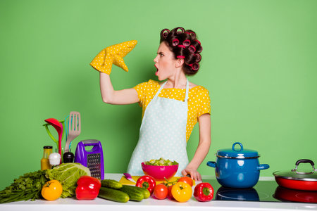 Profile side photo of shocked girl prepare tasty supper dish talk speak pot holder wear dotted yellow t-shirt hair rollers table cucumber tomato ingredients isolated green color backgroundの写真素材