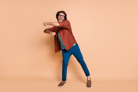 Full length photo of man dance raise fist crazy face wear eyewear brown t-shirt pants footwear isolated beige color backgroundの写真素材