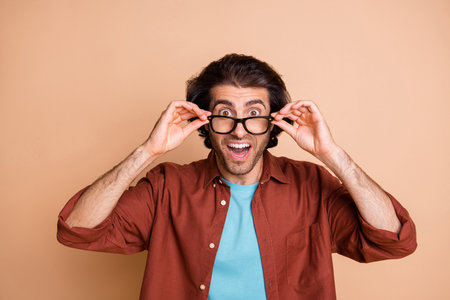 Photo of shocked young guy hands hold eyeglasses look camera wear specs brown t-shirt isolated beige...