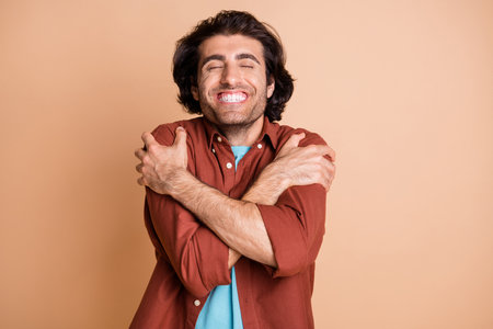 Photo of cheerful young man embrace shoulders eyes closed toothy smile wear brown t-shirt isolated...