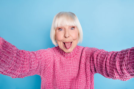 Photo portrait of funny elderly woman showing tongue taking selfie isolated on pastel blue colored...
