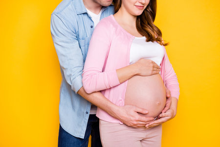 Cropped photo of young couple stand hug look pregnant woman stomach wear casual outfits isolated on yellow color backgroundの写真素材
