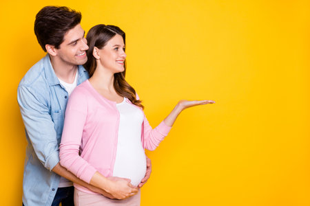 Profile photo of young couple stand hug pregnant woman hold empty space wear casual outfits isolated on yellow color backgroundの写真素材