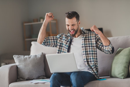 Photo of young excited man happy positive smile sit couch home look laptop celebrate win victory success fists handsの写真素材