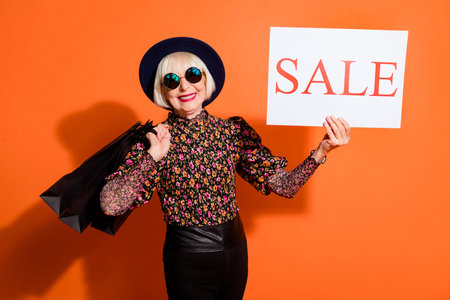Photo of happy cheerful smiling grandmother hold shopping and bags sale placard isolated on orange color backgroundの写真素材