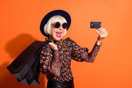 Photo of old excited happy smiling cheerful lady showing credit card hold shopping bags isolated on...