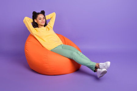 Full size photo of optimistic girl lying on pouf wear yellow shirt trousers sneakers isolated on lilac color backgroundの写真素材