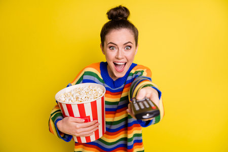 Portrait of pretty cute brunette girl eating pop corn hold remote controller wear rainbow sweater isolated on bright yellow backgroundの写真素材