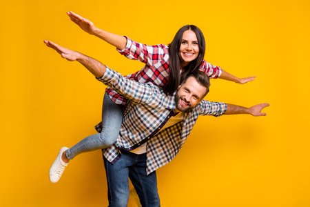 Portrait of nice lovely cheery couple piggy backing having fun like plane isolated over bright yellow color backgroundの写真素材