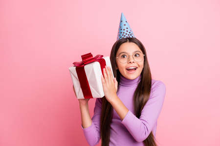 Photo of positive shocked small kid girl shake gift box wear violet birthday cap isolated over...