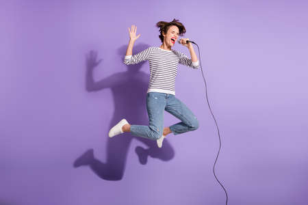 Full length photo of charming lady dressed casual clothes jumping singing microphone isolated purple color backgroundの写真素材