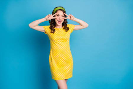 Photo of excited girl make v-sign near face wear green yellow polka-dot skirt isolated over blue color backgroundの写真素材