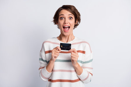Photo of impressed funny young lady wear striped sweater holding bank card isolated grey color...