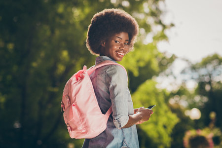 Rear view photo portrait of black skinned pretty girl keeping mobile phone walking in sunny park wearing pink backpack smilingの写真素材