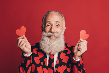 Photo of cheerful retired gentleman hold two little heart figures look camera wear suit isolated red backgroundの写真素材
