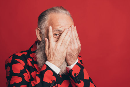 Photo of funny shy bearded man palms hide face peeking eye wear heart print suit isolated red backgroundの写真素材