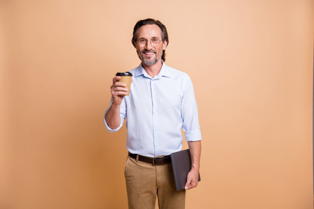 Portrait of nice cheerful man holding in hands laptop drinking take out latte isolated over beige...
