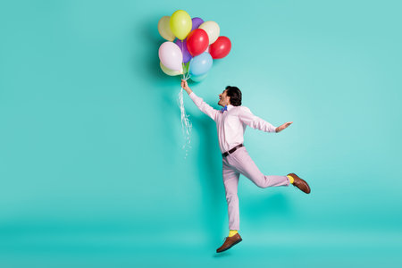 Portrait of jumping young man hold colorful helium balloons wear formal outfit yellow socks isolated on turquoise backgroundの写真素材