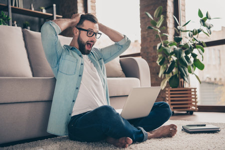 Full size profile side photo of happy amazed attractive man sit floor hold hand head look computer...