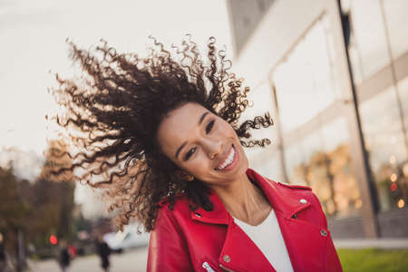 Photo of adorable sweet curly woman dressed red jacked walking street smiling air blowing curls sides outdoors urban city streetの写真素材