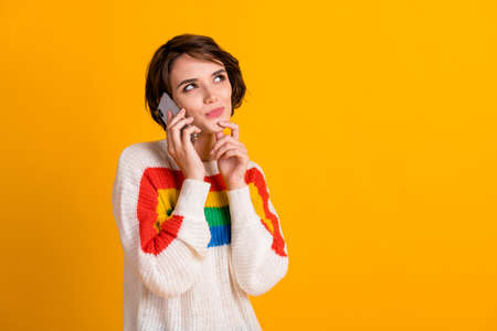 Photo of thoughtful girl hold smartphone hand chin look up empty space wear striped pullover isolated yellow color backgroundの写真素材