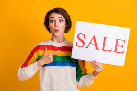 Photo of surprised pretty brown short hair lady stand hold placard sale point finger wear rainbow sweater isolated yellow color backgroundの写真素材
