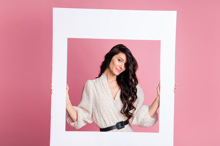 Photo portrait of cheerful woman keeping photo frame wearing summer dotted white dress isolated pastel pink color backgroundの写真素材