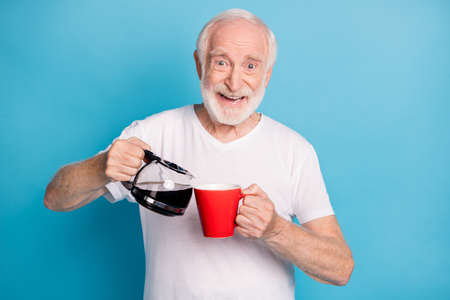 Photo portrait of retired man pouring coffee in red mug isolated on pastel blue colored background