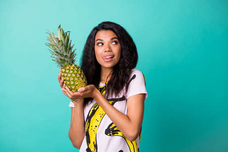 Photo of dark skin girl hold pineapple look empty space lick lips tongue wear pink t-shirt isolated...