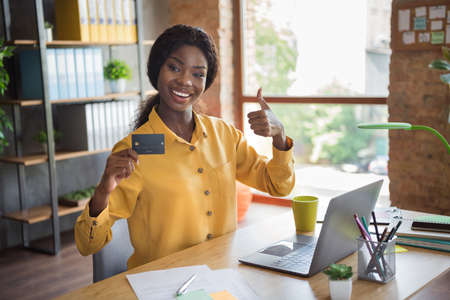 Photo portrait of african american woman holding credit card showing thumb up in modern office indoorsの写真素材