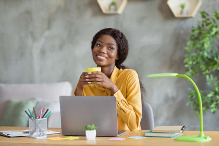 Photo of happy dreamy dark skin young business woman sit office desk relax drink coffee job indoors workstationの写真素材