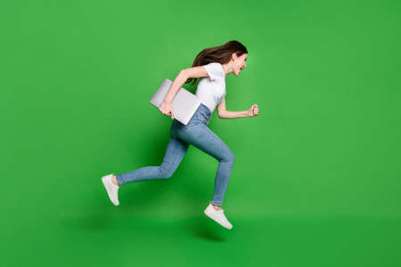 Full length profile side photo of crazy girl hold laptop jump run wear t-shirt denim isolated green color backgroundの写真素材