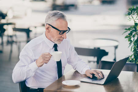 Profile photo of optimistic grey hair old business man drink tea write laptop wear spectacles white shirt outdoors near work centerの写真素材