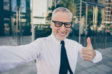 Photo of optimistic grey hair old business man show thumb up do selfie wear spectacles white shirt outdoors near work centerの写真素材