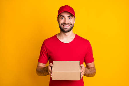 Photo portrait of delivery worker wearing red cap t-shirt keeping box for packing isolated on vivid yellow color backgroundの写真素材