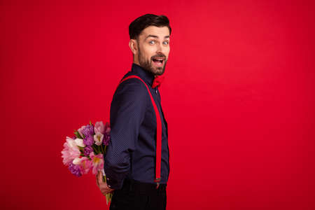 Profile side photo of young good mood excited amazed gentleman hide bouquet surprise isolated on red...
