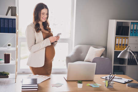 Photo portrait of pregnant woman holding phone in two hands typing standing in modern officeの写真素材