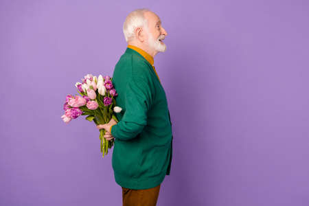 Profile photo of neat aged man look empty space hands hold flowers behind back isolated on violet...