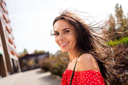Photo of cute charming young lady wear red off-shoulders dress wind blow hair smiling walking outside urban city streetの写真素材