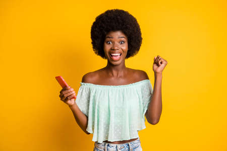 Photo portrait of woman celebrating with fist up holding phone in one hand isolated on vivid yellow colored backgroundの写真素材