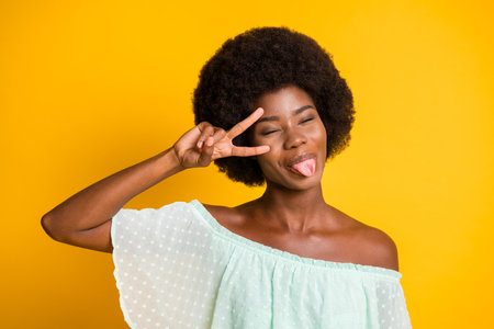 Photo portrait of girl showing tongue holding v-sign near eye isolated on vivid yellow colored backgroundの写真素材