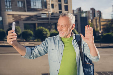 Photo of friendly cute age gentleman wear denim shirt backpack recording video modern gadget waving arm outdoors city streetの写真素材