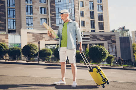Photo of cute attentive age guy wear denim shirt cap walking holding luggage looking map outdoors street urban cityの写真素材